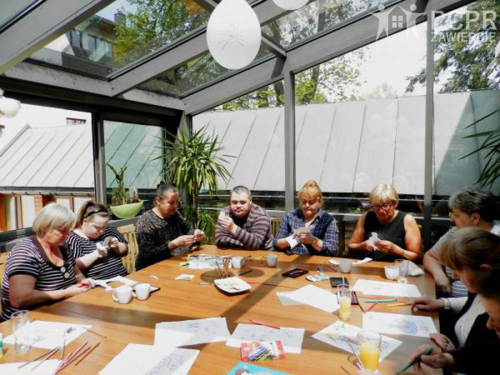 Zdjęcie: Grupa kobiet i mężczyzn, siedząc przy stole, wykonuje wycinanki z kartki papieru
